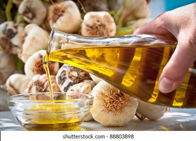 Hand Pouring Extra-virgin Olive Oil In A Bowl With Fresh Garlic In The Background.