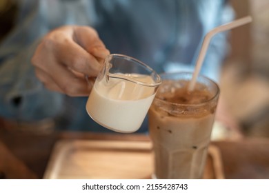 A hand pouring a glass of milk cream into iced latte coffee on a wooden bar over a cafe glass window reflex at a Cafe coffee shop. Cold brew refreshment summer drink with copy space. Selective focus.