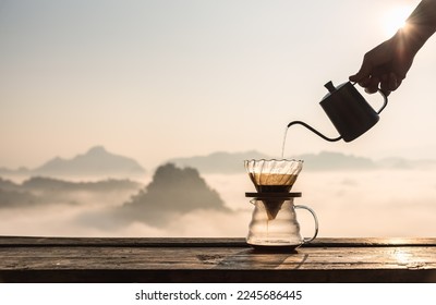 Hand pouring hot water on coffee ground with fillter, barista using drip coffee kit set with mountain and fog in sunrise background. - Shutterstock ID 2245686445