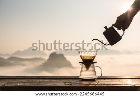 Hand pouring hot water on coffee ground with fillter, barista using drip coffee kit set with mountain and fog in sunrise background.
