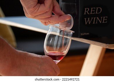 Hand pouring red wine into a glass from a BIB - cardboard bag in box with open tap standing on a table. Close up image. Fictitious brand.