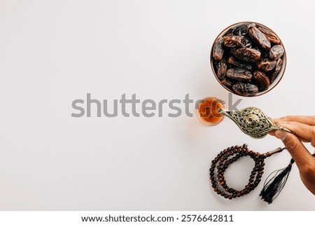 Hand pouring tea from a teapot with dates and prayer beads on white. Ramadan celebration highlights holy traditions with a serene flat lay arrangement featuring tea and unity.