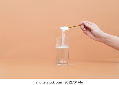 Hand pours collagen protein powder in a glass of water on a beige background. A natural supplement for skin beauty and bone health. Space for text. - Shutterstock ID 2012704946