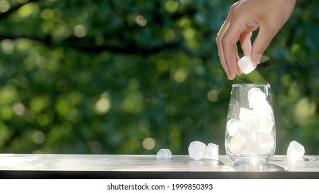 Hand pours many ice cubes into a transparent glass. Making drinks outdoors. Glass on the brown organic wooden table with green morning summer trees leaves bokeh background