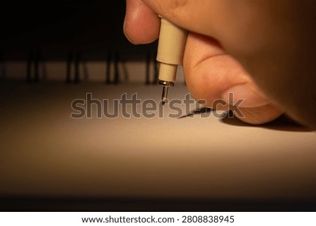 A hand preparing to write or draw on a notebook page with a fine liner pen, illuminated by warm spotlighting, ready for creation