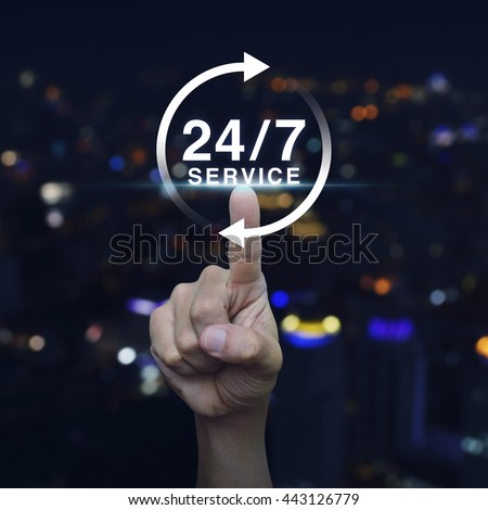 Hand pressing button 24 hours service icon over blurred light city tower background, Full time service concept