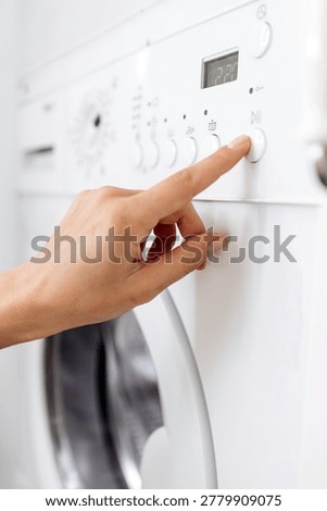 Hand pressing the button on a white washing machine with a digital display and control panel in a clean laundry room setting. Vertical shot
