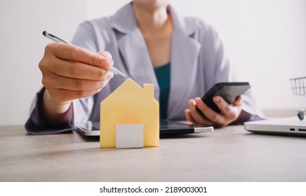 The Hand Is Pressing Calculators, Piggy Bank With Wooden House. Buy Or Rent Question On Note With Calculators On Desk. Save Money And Buy House Concept.