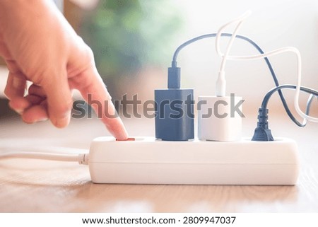 Hand pressing the red switch on a power strip, disconnecting multiple chargers and plugs to reduce electricity consumption and promote energy efficiency