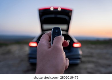 Hand pressing trunk button on car key fob in front of vehicle with open trunk and illuminated rear lights at dusk.
