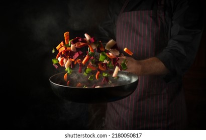 The hand of a professional chef throws pieces of vegetables into a hot frying pan with steam on a black background. The concept of cooking in the hotel. Free ad space - Shutterstock ID 2180050817