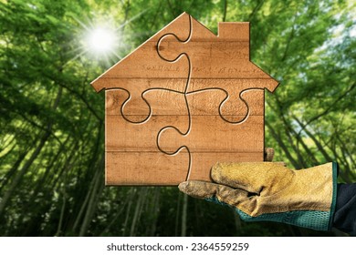 Hand with protective work glove holding a small wooden house made of jigsaw puzzle pieces with a green forest on background and sun rays. Eco house in green environment concept. - Shutterstock ID 2364559259