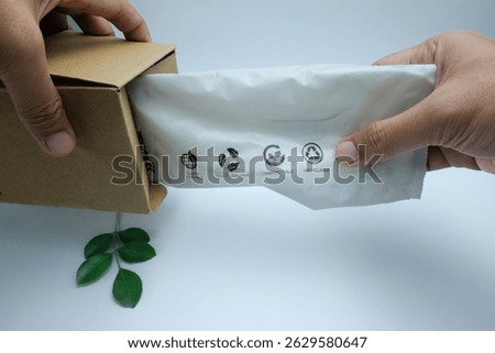 A hand pulls out a biodegradable plastic bag from a cardboard box. The bag features clear recycling, compostable, and biodegradable symbols. Illustrates sustainable packaging, environmental awareness