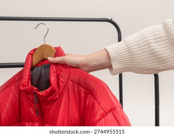 Hand pulls red puffer jacket from metal rack against white background. Quilted outerwear hangs on wooden hanger along black rail. Minimalist retail fashion concept for wardrobe choice in boutique shop