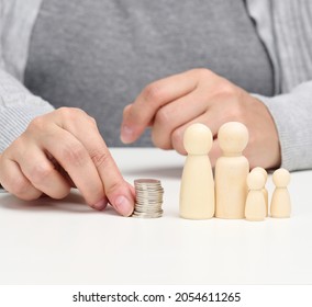 Hand Puts Coins In A Pile And Wooden Figurines Of A Family On A White Table. Economy And Investment Concept, Budget Planning. Receiving Subsidies From The State, Tax