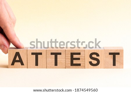 The hand puts a wooden cube with the letter A from the word Attest. The word is written on wooden cubes standing on the yellow surface of the table.