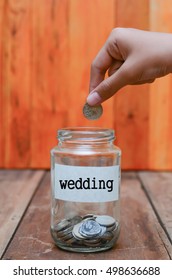 Hand putting a coin into a glass jar with wooden table and background with word WEDDING on it.

 - Shutterstock ID 498636688