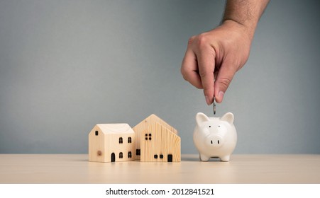 Hand Putting A Coin In Piggy Bank Around With Wooden House Model On Gray Background. Saving Money For Home Buying Concept.