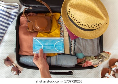 Hand Putting A Face Mask On The Suitcase Prepared To Travel In Summer, In The New Normal, After The Coronavirus Covid 19 Pandemic. Concept Of Quarantine, Coronavirus, Summer Vacation In New Normal.