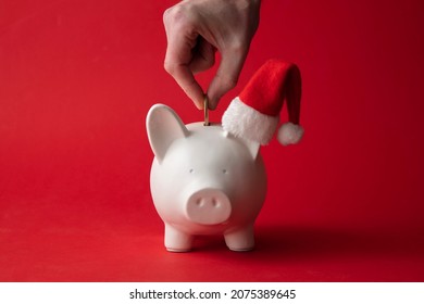 Hand Putting Money Into A Piggy Bank Wearing A Santa Hat. Christmas Savings Background