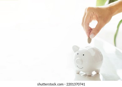 Hand Putting Money In White Piggy Bank With Tiny House On White Marble Background And Blurred Bokeh With Copy Space. Concept For Financial Home Loan Or Money Saving For House Buying, Real Estate And