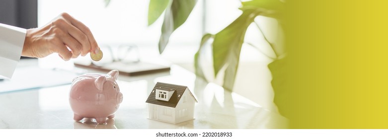 Hand Putting Money In White Piggy Bank With Tiny House On White Marble Background And Blurred Bokeh With Copy Space. Concept For Financial Home Loan Or Money Saving For House Buying, Real Estate And