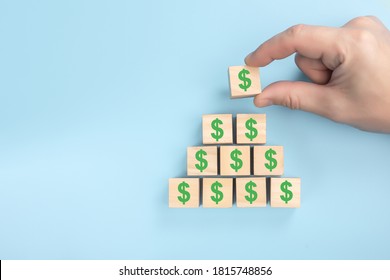 Hand Putting Wooden Cube Block On Top Pyramid Of Dollars. Wood Block Cubic Stacking Up With Green Dollar Sign. Blue Background. Business Growth Success Process. Investment Concept