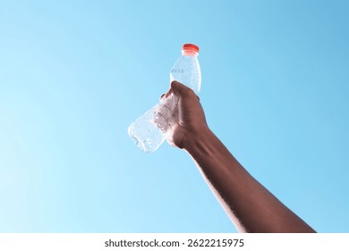 A hand raises an empty plastic bottle against a clear blue sky, symbolizing hope and a call for environmental awareness. This image represents the importance of recycling, reducing plastic waste