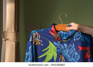 ็็Housewife's Hand Raising Her Husband's Hawaiian Shirt On Hanger To Check For Tidy And Cleanliness On Green Wall At Home