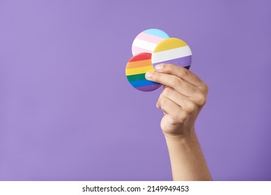 Hand raising three pins with rainbow, trans and non binary flags, on a purple background. Concepts of LGBT identity pride, gender diversity visibility, equality and non discrimination.