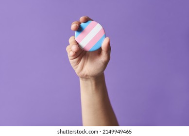 Hand raising a trans flag badge or pin on a purple background. Concepts of identity pride, gender diversity visibility, equality and non discrimination. - Shutterstock ID 2149949465