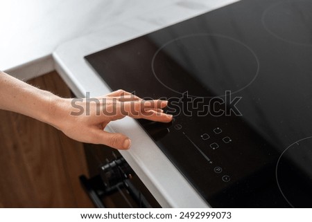 A hand reaches down to adjust the temperature of a black electric stove top burner. Woman press start button on touch screen of cooker in kitchen, close up shot