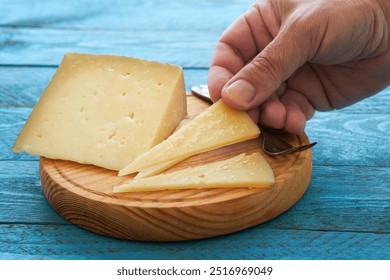 A person’s hand reaches for a slice of artisan cheese on a wooden board, surrounded by metal utensils, highlighting the texture and color against a vibrant blue surface. - Powered by Shutterstock