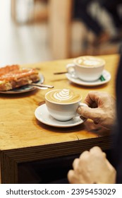Hand reaching for a cappuccino with latte art on a wooden cafe table, another cup and plate in the background - Shutterstock ID 2395453029