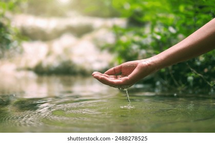 hand is reaching into a body of water, splashing water out of its fingers. The scene is peaceful and calming, with the water reflecting the sunlight and the surrounding greenery - Powered by Shutterstock