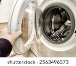 Hand reaching for the open door of a modern washer in a cozy laundry area while completing household chores.