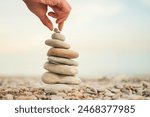 A hand is reaching for a stack of rocks, which are arranged in a pyramid shape. Concept of balance and harmony, as the rocks are stacked in a way that creates a visually pleasing and stable structure