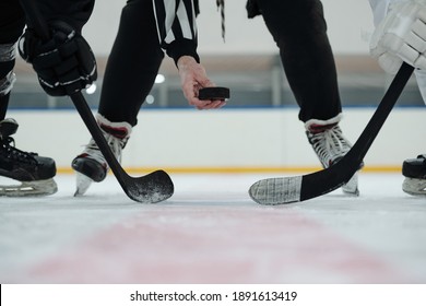 Hand of referee holding puck over ice rink with two players with sticks standing around and waiting for moment to shoot it