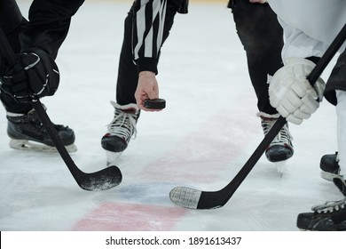 Hand of referee holding puck over ice rink with two hockey players with sticks standing around him and waiting for moment to shoot it