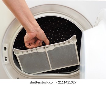 Woman’s hand removing fluff filter into the tumble dryer of clothes dryer with blurred washed clothing inside. Laundry processes, cleaning and care concept, personal hygiene theme. (selective focus)
