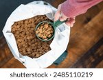 Hand removing a green measuring cup full of dog or cat food kibble from a white bag full of round pieces of food.