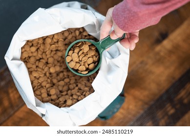 Hand removing a green measuring cup full of dog or cat food kibble from a white bag full of round pieces of food.