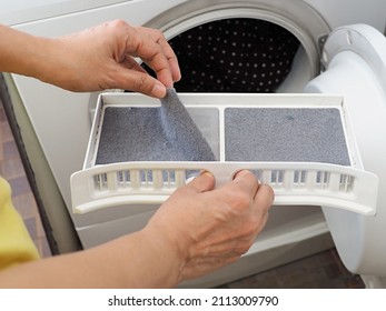 Person’s hand removing lint from fluff filter of the tumble dryer on blurred background of clothes dryer with washed clothing. Laundry processes, Cleaning and care concept. (close up, selective focus)