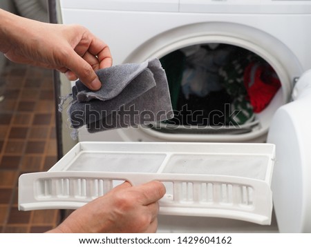 Woman’s hand removing lint from fluff filter of the tumble dryer on blurred background of clothes dryer with washed clothing. Laundry processes, Cleaning and care concept. (close up, selective focus)