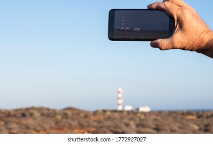 Hand of a retired elderly man while using his cellphone to photograph the lighthouse in the distance.