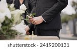 Hand, rose and a person at a funeral in a graveyard in grief while mourning loss at a memorial service. Death, flower and an adult in a suit at a cemetery with a coffin for an outdoor burial closeup