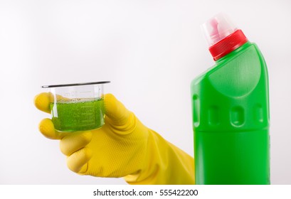 Hand With Rubber Glove Holding Dosage Cup With Green Detergent And Bottle Of Cleaning Product Against White Background