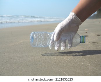 Hand In Rubber Gloves Picking Up Clear Plastic Water Bottle Raised On A Beach By The Sea Against Background Of Sea Water And Sky Or Concept Garbage On The Beach