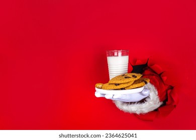 Hand of Santa Claus with traditional Christmas cookie snack with milk glass. Santa Dunking Cookie, on bright festive red torn paper hole background. Merry Christmas and Happy New Year greeting card - Shutterstock ID 2364625041