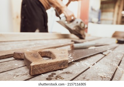 Hand saw with a wooden handle on blur carpenter working with electric wood planer. Carpenter tools. Hand saw and sawdust on wood table at outdoor workshop. Craftsman making woodwork. Handwork concept.